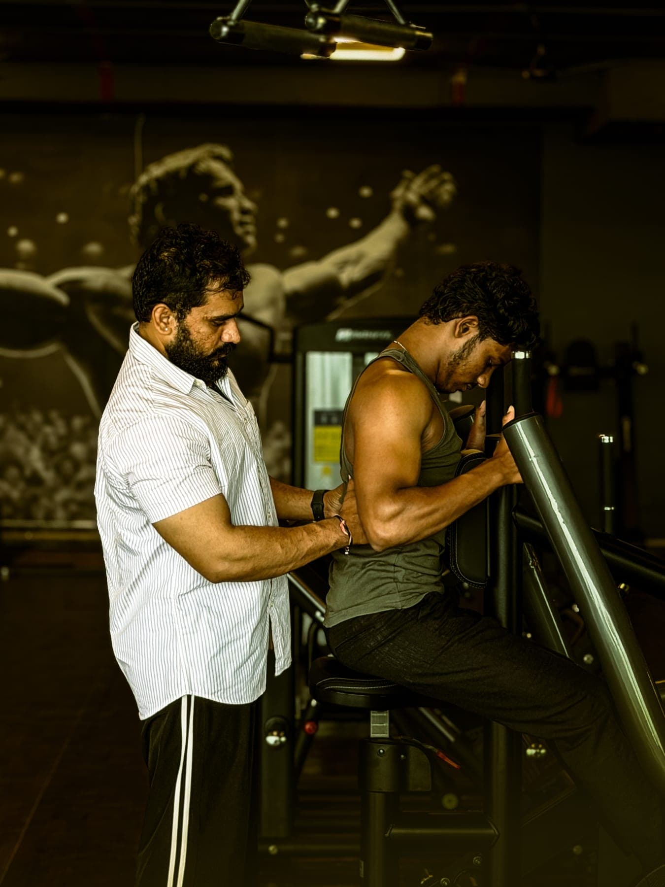 Member training on the shoulder press machine with trainer guidance, beneath the gym's mural